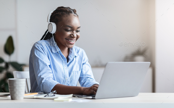Smiling black secretary working on laptop and listening music in headphones at office, sitting at desk on workplace, free space