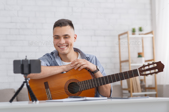 Online music lesson. Smiling guy with guitar looks at smartphone camera in living room interior, free space