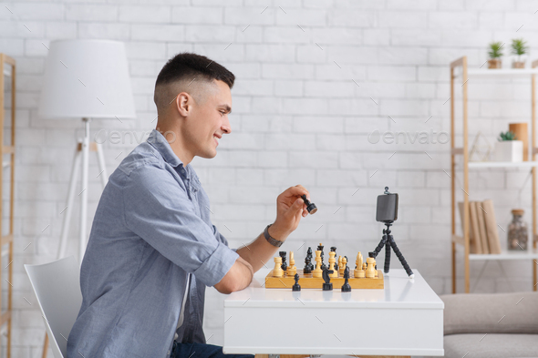 Online hobby at home. Smiling young man in jeans shirt plays chess at table  Stock Photo by Prostock-studio