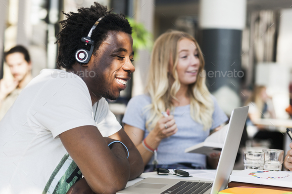 Happy male college student listening music through headphones while using laptop in cafe