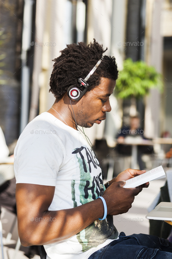 Side view of male college student listening music while reading book at cafe