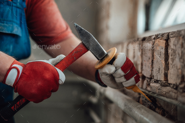 Worker’s strong hands with hammer and chisel, getting rid of plaster on a brick wall