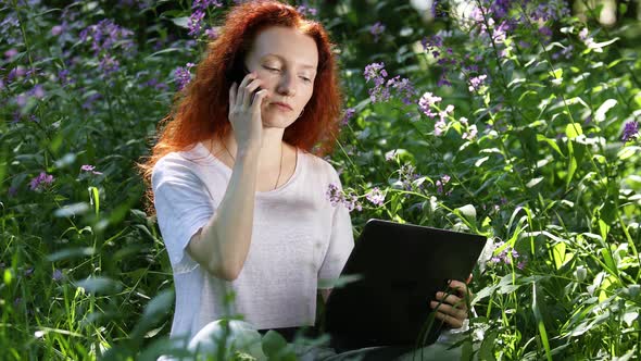 Young Red-Haired Woman Work On Laptop Computer And Talking On a Mobile Phone Among Flowers