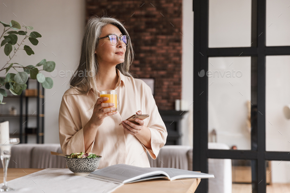 Photo of mature amazing concentrated gorgeous woman at kitchen indoors at home chatting by phone and drinking juice.