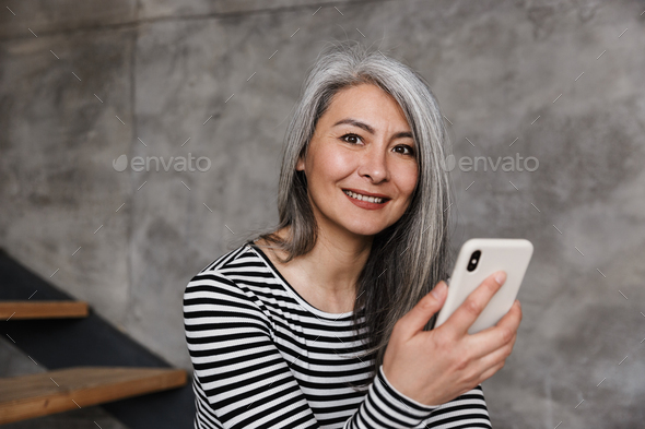 Photo of happy adult asian woman smiling and using cellphone while sitting on stair at home