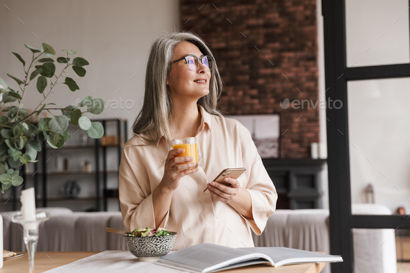 Photo of mature amazing positive dreaming gorgeous woman at kitchen indoors at home chatting by phone and drinking juice.