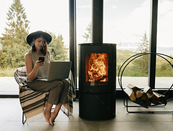 Stylish hipster girl sitting with laptop on cozy chair near fireplace with fire - Stock Photo - Images