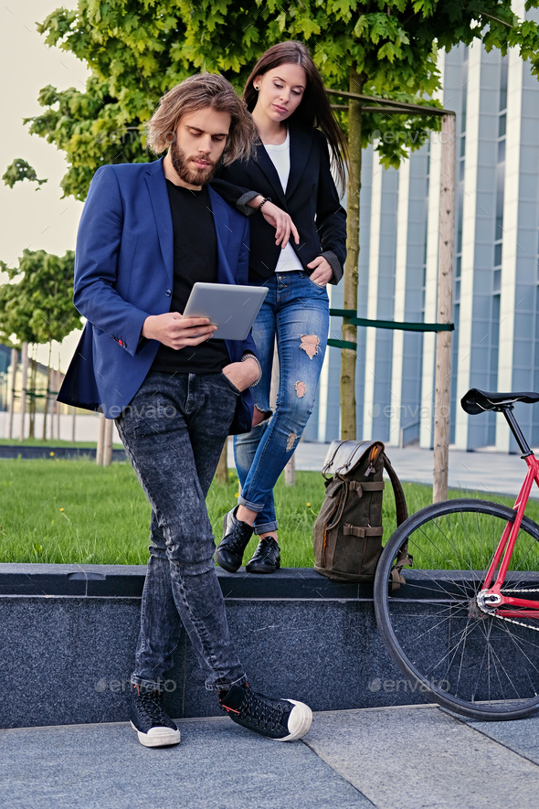 Stylish urban couple using a tablet PC with fixed red bicycle in a park on background.