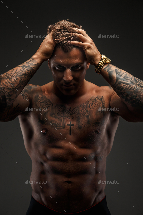 Shirtless muscular tattooed man posing in shadow over grey background.