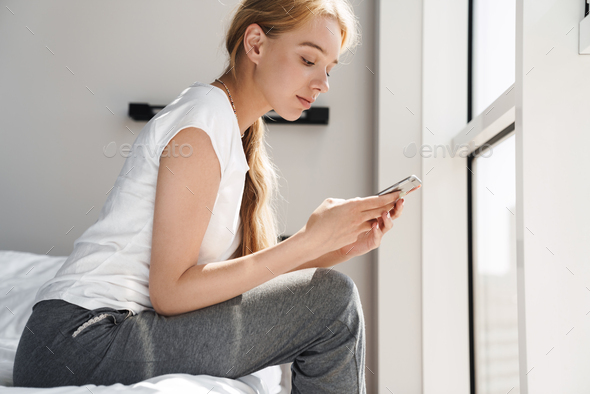 Photo of focused young woman using cellphone while sitting in bed after sleep at bright room