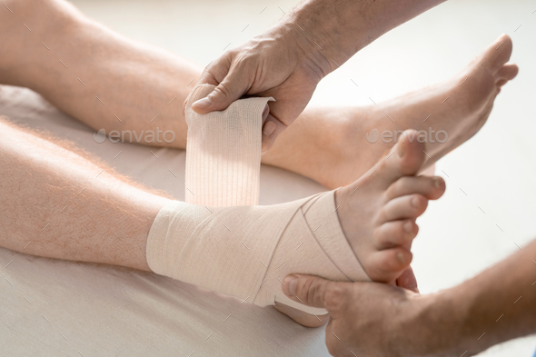 Hands of rehabilitation clinician wrapping foot and ankle of patient with flexible bandage during medical procedure on couch in hospital