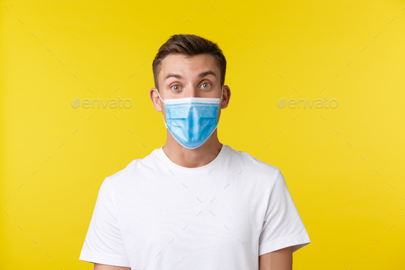Concept of social distancing, covid-19 and people emotions. Excited and surprised handsome guy found out awesome news, wearing medical mask, looking amazed over yellow background.