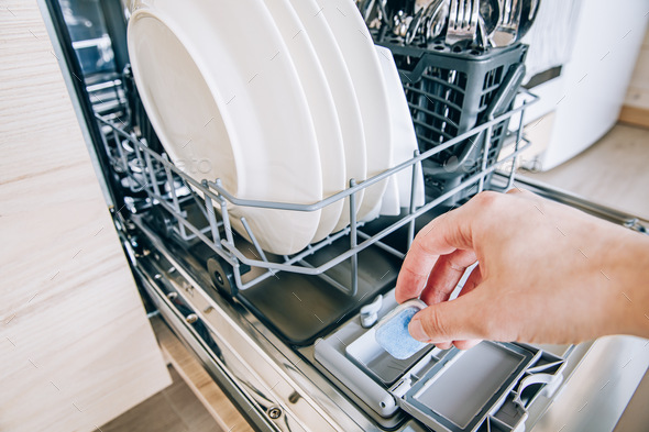 Woman hand putting tablet in dishwasher detergent box. Dishwasher machine full loaded.