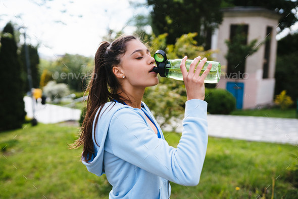 Young woman in a sporting suit drinks water from a bottle after outdoor gymnastics in the summer