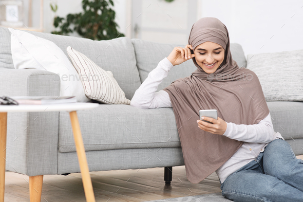 Millennial Arabic Girl In Headscarf Using Smartphone At Home, Texting With Friends, Sitting On Floor In Living Room, Free Space