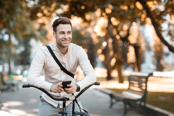 Street style. Young stylish man texting on phone while sitting on bike in park, free space