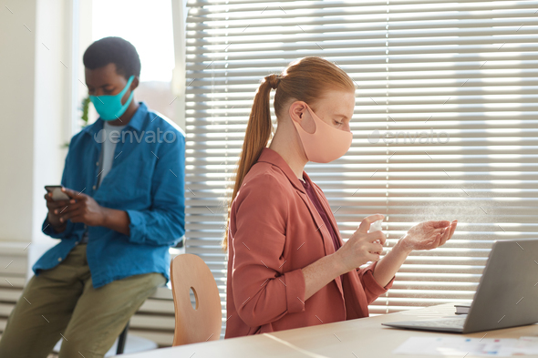 Side view portrait of young woman wearing face mask sanitizing hands at workplace in post pandemic office, copy space