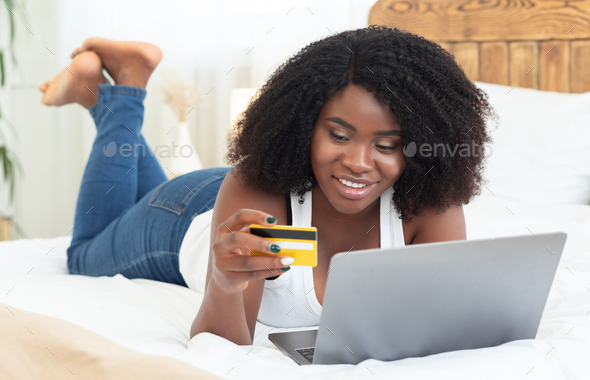 Lovely black girl lying on bed at home with laptop computer and credit card, shopping online