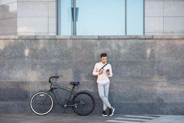 Online outdoors. Businessman with bicycle and cup of coffee, looks at smartphone leaning against the wall during break from work in city, copy space