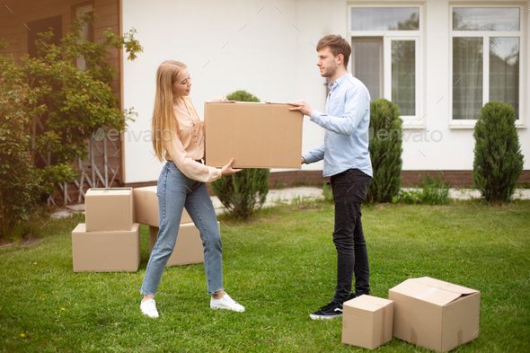 Young married couple carrying cardboard box in front of their new house on moving day