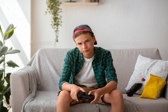 Bored teen boy holding joystick, sitting on couch in his room, eating junk food, copy space