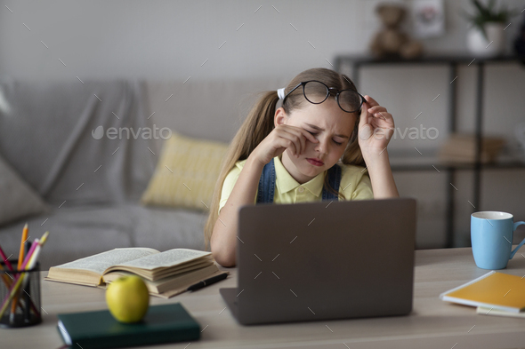 Tired Student. Portrait of kid taking off glasses, touching dry eyes, sitting at table working on laptop