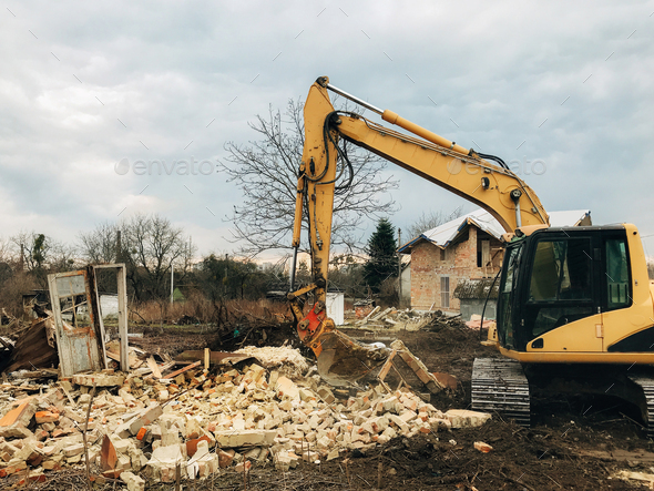 Bulldozer clearing land from old bricks and concrete from walls with dirt and trash. Backhoe machinery ruining house. Excavator destroying brick house on land in countryside.