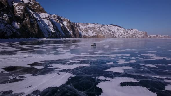 Flying of a Drone Over the Ice of the Lake Behind a Driving Car Along the Rocky Coast alt
