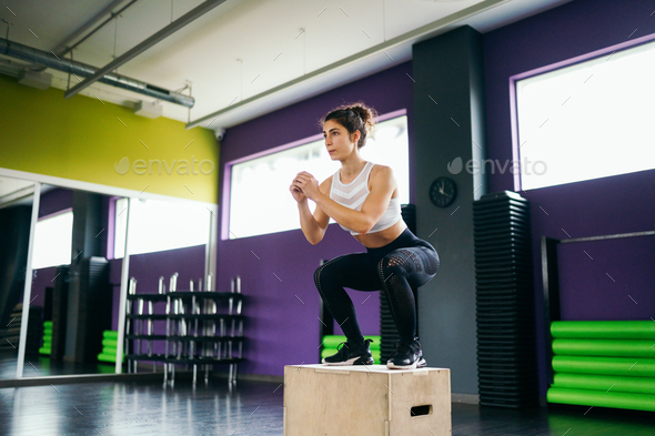 Athletic woman doing squats on box as part of exercise routine. Caucasian female doing box jump workout at gym.