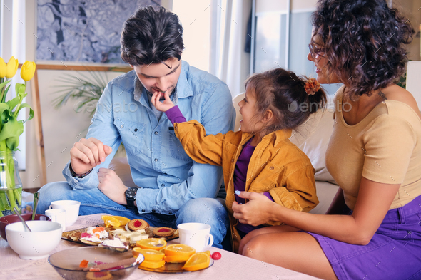 Attractive brunette female in eyeglasses, bearded stylish male and their cute little daughter sits on a couch and eat lunch in a living room.