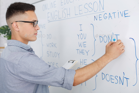 Online school and teacher during quarantine. Serious man in glasses writes rules of english on white board in living room interior, side view, close up, copy space