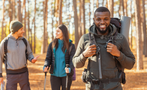 Happy black guy hiking with caucasian friends by countryside, forest background