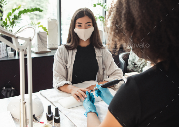Work of manicure and pedicure salon during covid-19. Asian girl looks at african american master in protective mask during electrical nail procedure in studio interior