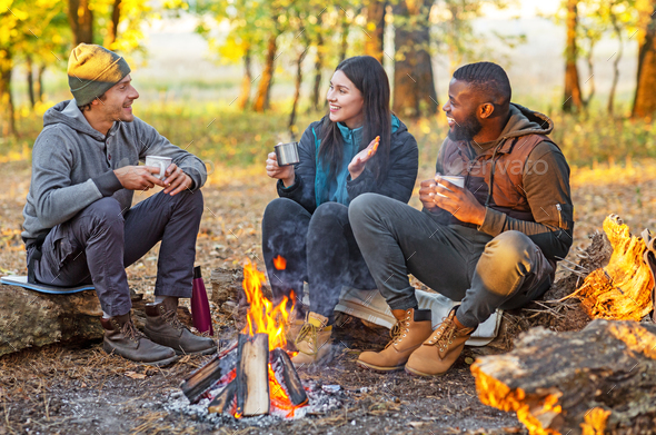 Group of closed multiracial friends hikers enjoying hot tea next to camp fire in sunset lights