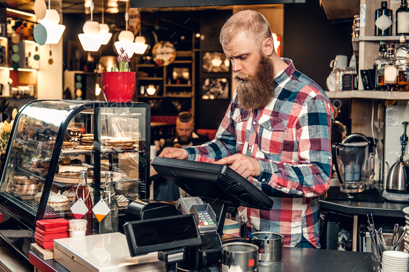 Positive bearded male at the counter using cash register in a coffee shop.