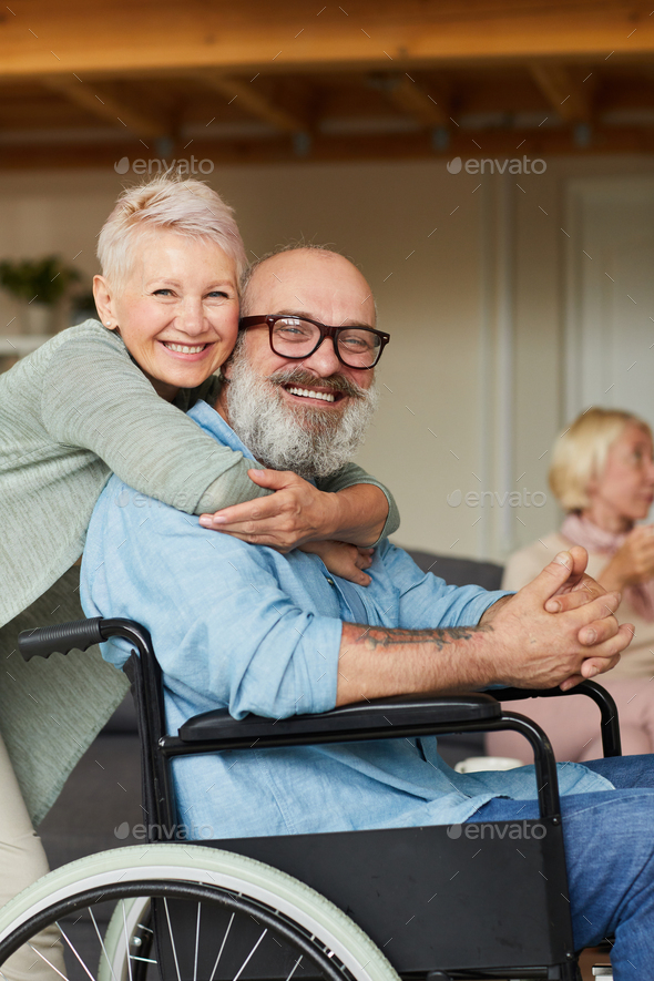 Portrait of happy senior woman embracing her disabled husband and they smiling at camera
