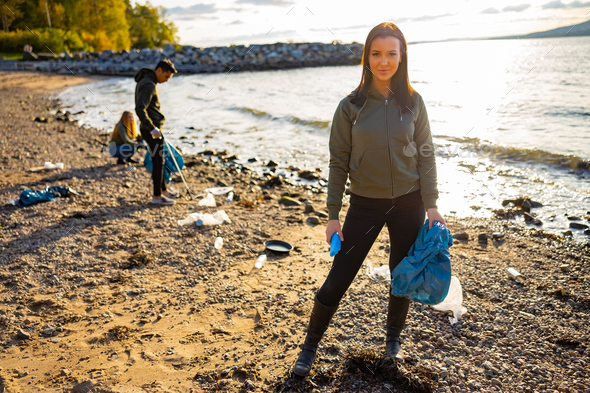 Smiling young woman cleaning beach with volunteers during sunset - Stock Photo - Images