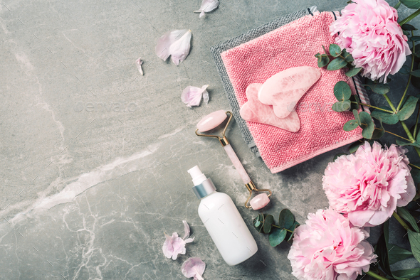 Cosmetic products bottles, towels, oil, face roller and gua sha massager, pink peonies flowers on