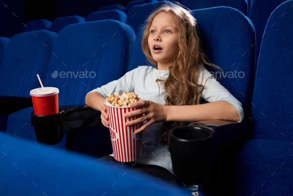 Side view of excited female teen watching action film in cinema. Little girl holding popcorn and sweet water, having rest and relaxing during weekend. Concept of childhood, entertainment.