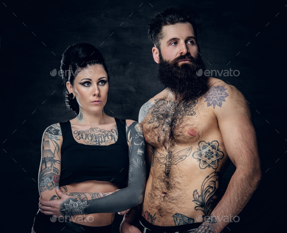 Portrait of shirtless, tattooed bearded hipster male and brunette female with tattoo ink on her torso over dark grey background.