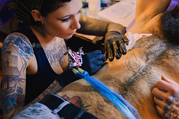 A professional female tattoo artist makes black and white tattoo on bearded male torso in a salon.