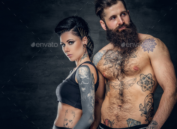 Portrait of shirtless, tattooed bearded hipster male and brunette female with tattoo ink on her torso over dark grey background.