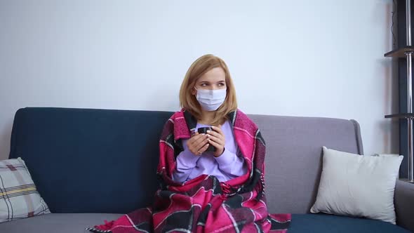 The Girl Is Sick Sitting at Home on the Sofa in a Protective Mask in Her Hands Holds a Cup alt