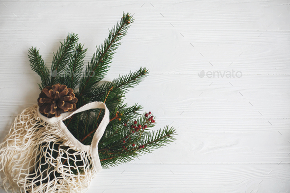 Net shopping bag with winter decorations, zero waste holidays. Top view with space for text. Reusable cotton bag with green spruce branches and pine cones on white rustic background.