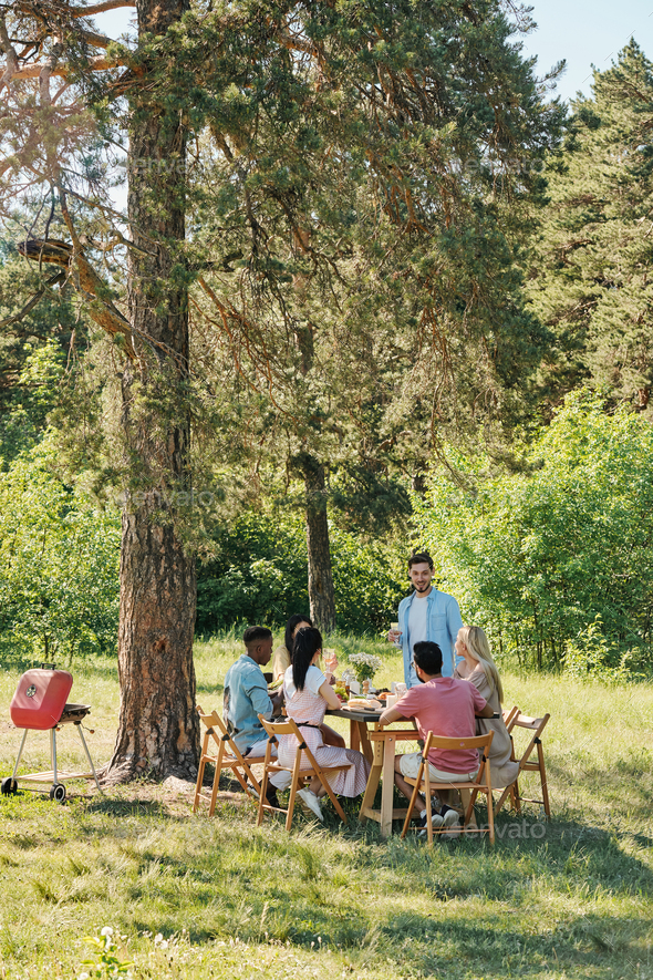 Young man standing by served table in front of his friends during outdoor dinner under pine tree and pronouncing toast on summer weekend