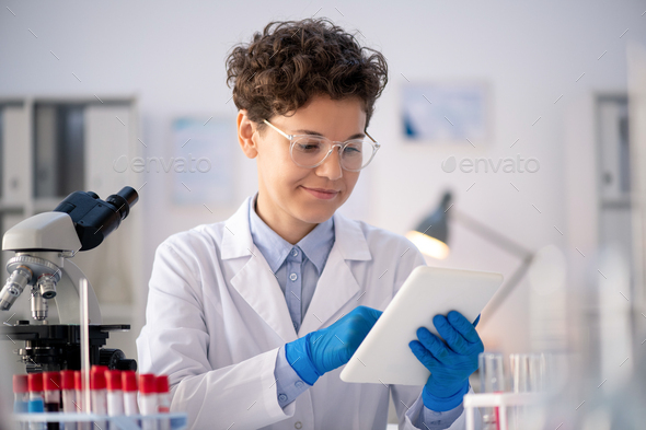 Happy young contemporary female scientist or pharmacist in gloves, whitecoat and eyeglasses looking through online information