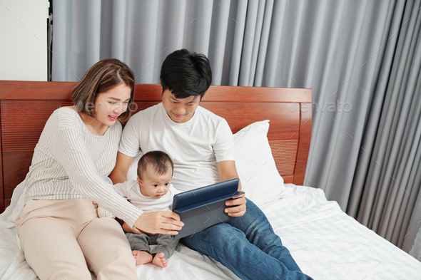 Parents and little kid watching interesting cartoon on tablet computer when resting on bed in the evening