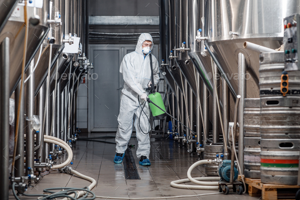Factory cleaning. Man in protective suit and mask disinfects plant and brewing kettles during covid-19