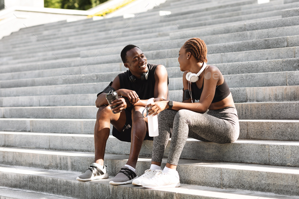 Positive African Couple Resting After Fitness Workout Outdoors, Sitting On Urban Steps, Drinking Water And Chatting, Copy Space