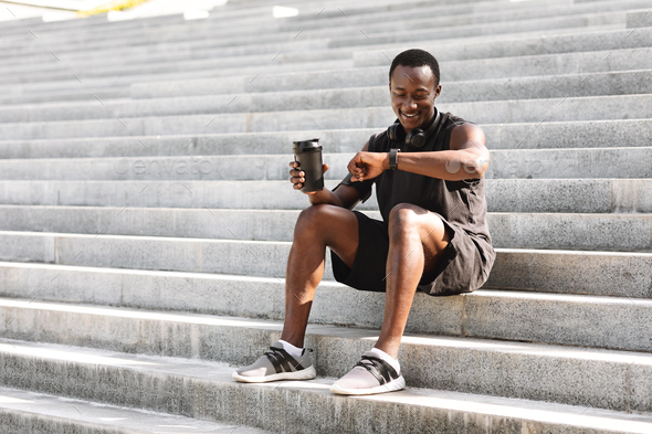 Smiling African Athete Guy Checking Fitness Tracker After Training Outdoors, Sitting On Urban Steps, Copy Space